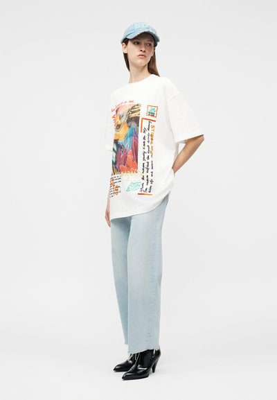 Γυναίκα που φοράει φαρδιά λευκή μπλούζα με σχέδιο, ανοιχτό μπλε τζιν παντελόνι φαρδιάς γραμμής, μαύρα μπότες με τακούνι και ανοιχτό μπλε καπέλο μπέιζμπολ, στέκεται με τα χέρια στις τσέπες.