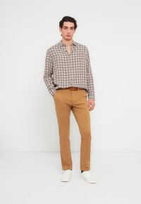 Καφέ και λευκό πουκάμισο με καρό μοτίβο, ανοιχτό γιακά, σε συνδυασμό με εφαρμοστά μπεζ παντελόνια. Λευκά αθλητικά παπούτσια. Ομαλή υφή και casual σχεδιαστικά στοιχεία.