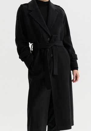 Person trägt einen langen schwarzen Trenchcoat mit Gürtel, mit geknöpften Riemen an Ärmeln und Schultern, steht mit einer Hand in der Tasche.