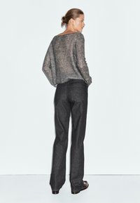 Donna in piedi con le mani in tasca, indossando un maglione grigio a maglia larga e jeans dritti scuri, su uno sfondo semplice chiaro.