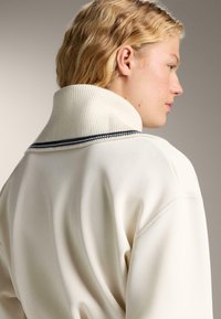 Tânăr cu păr blond scurt și ondulat, îmbrăcat cu un pulover alb cu un guler înalt cu nervuri, având o dungi subțire neagră.