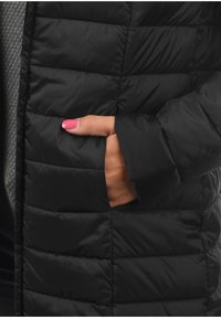 Černá péřová bunda s prošitou texturou a boční kapsou. Detailní záběr ukazuje hladkou tkaninu a ruku spočívající v kapse.