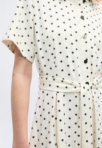 Λευκό φόρεμα με κοντά μανίκια και μαύρα πουά, με κουμπιά μπροστά και ζώνη από το ίδιο ύφασμα δένεται στη μέση.