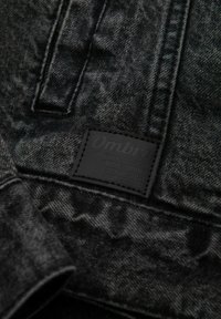 Tessuto denim nero con una finitura testurizzata. Presenta un'etichetta rettangolare in cuoio nero con la scritta "Ombre" e dettagli di cucitura visibili.