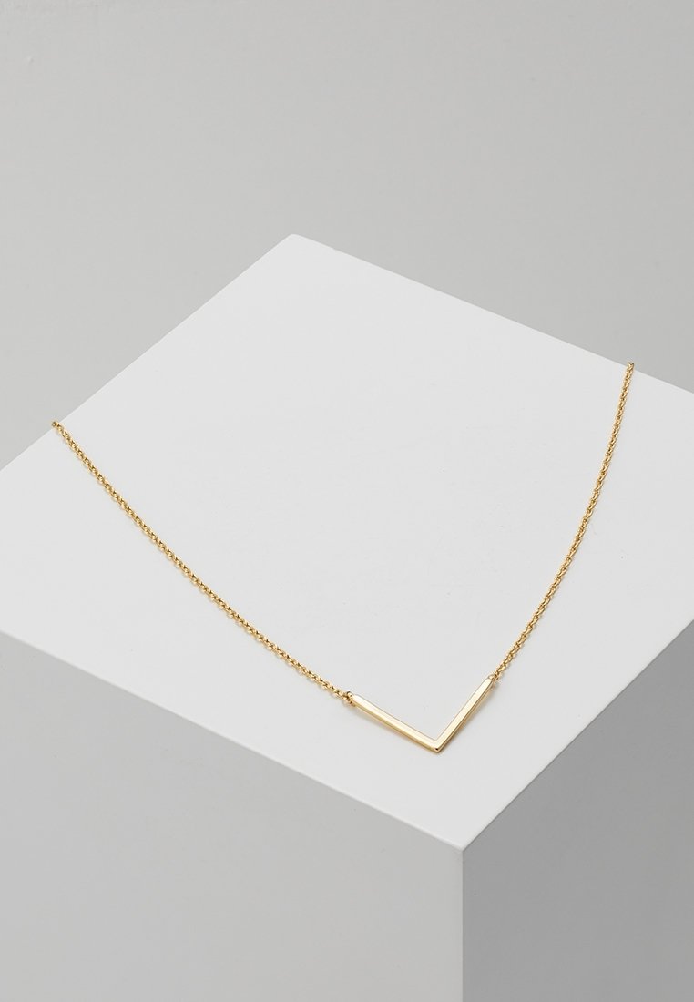 Collier délicat en chaîne d'or avec un petit pendentif en forme de V présenté sur une surface géométrique blanche sur fond gris.