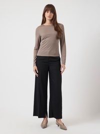 Nő egy sima háttér előtt áll, taupe színű, hosszú ujjú felsőt, fekete, széles szárú nadrágot elöl hasítékkal és taupe színű, alacsony sarkú cipőt visel.