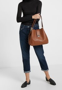 Woman dressed in black long-sleeve top and cuffed blue jeans holding a large brown leather handbag with gold hardware.
