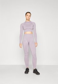 Ljubičasti sportski crop top s dugim rukavima i odgovarajuće visoko strukirane tajice, izrađene od rastezljivog materijala, u kombinaciji s crnim tenisicama.