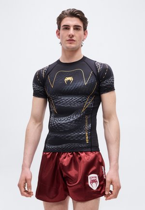 Młody mężczyzna w czarnej, przylegającej koszulce z motywem węża i żółtym logo oraz czerwonych satynowych szortach sportowych z białym logo, stojący na białym tle.