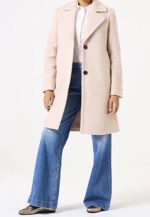 Personne portant un manteau en laine beige sur une chemise blanche, un jean bleu à jambes larges et des chaussures métalliques pointues, debout devant un fond uni.