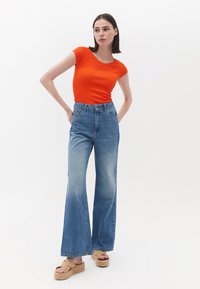 T-shirt aderente arancione abbinato a jeans a vita alta con fondo svasato di un lavaggio chiaro. Il modello indossa sandali con tacco a cuneo beige, in piedi contro uno sfondo bianco.