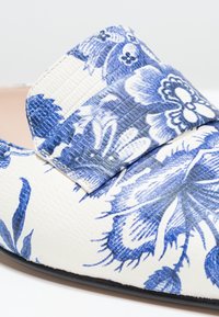 Chaussure blanche avec motif floral bleu et surface texturée en gros plan, montrant les détails avant et latéraux.