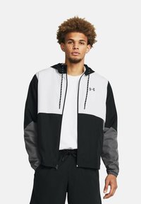 Chaqueta capucha en blanco y negro con detalles grises, que cuenta con cordones ajustables, cierre frontal con cremallera y puños elásticos. Llevada sobre una camisa blanca.