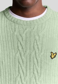 Maglione a coste di colore verde chiaro con un colletto a coste e un piccolo logo giallo e nero in basso a destra.