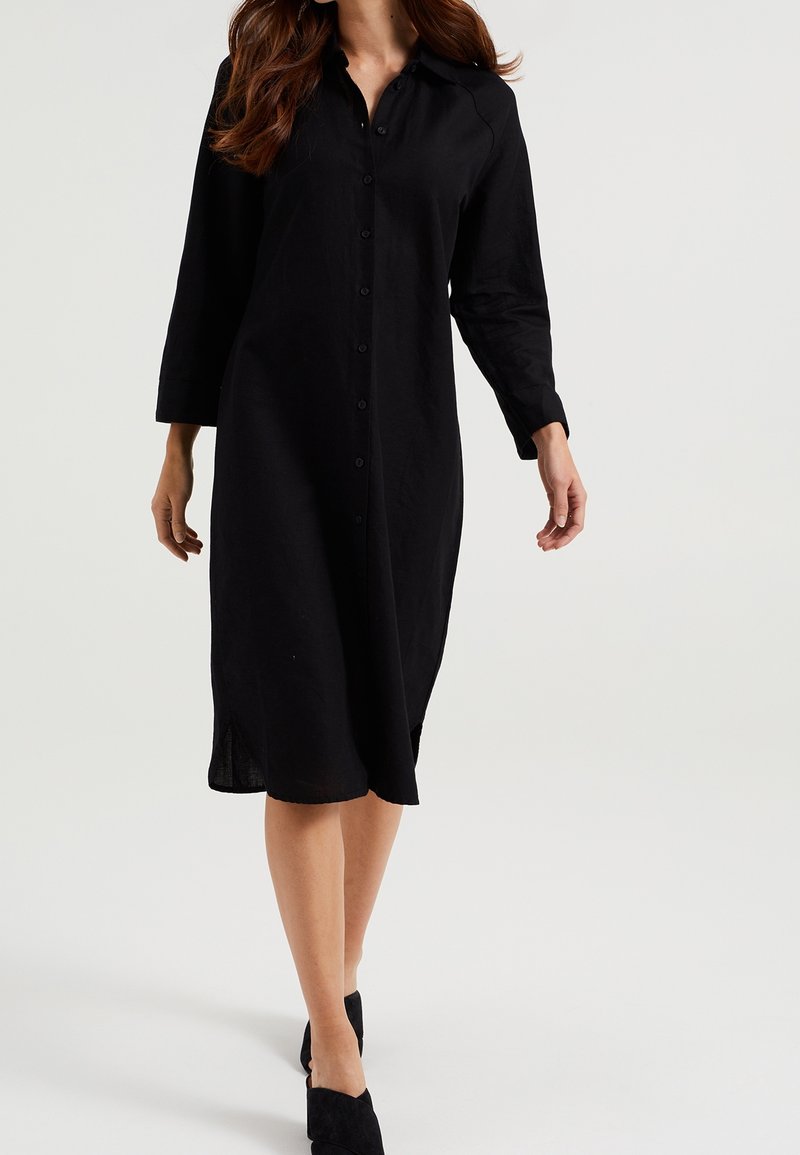 Svart skjortklänning gjord av lättviktigt tyg, med långa ärmar, krage, sidofåror och design som går till knäna.