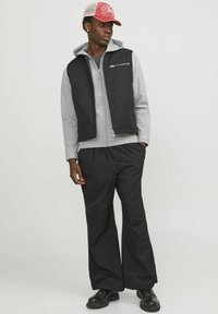 Chaleco sin mangas negro sobre una sudadera gris con cremallera, combinado con pantalones sueltos negros y botas negras. La gorra es roja con una parte trasera de malla gris.