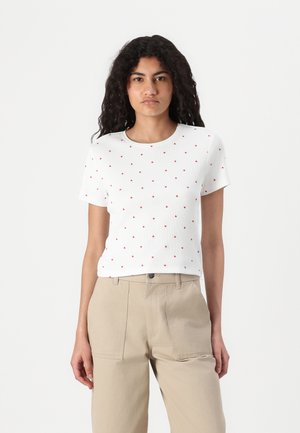 Witte t-shirt met korte mouwen en rode hartpatronen. Ribbelstructuur, ronde halslijn en een relaxte pasvorm. Gecombineerd met beige broek.