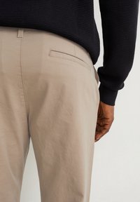 Personne portant un pantalon beige et un pull en maille noir, vue de dos, avec une main reposant le long du corps.