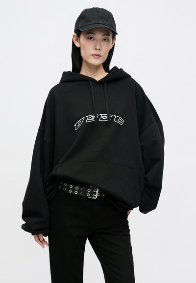 Fekete oversized kapucnis pulcsi elülső logóval, állítható kapucnival és széles ujjakkal. Fekete nadrággal és szegecses övvel párosítva.