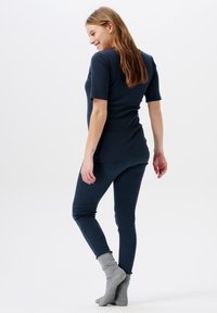 Vrouw die glimlacht, op haar tenen staat, gekleed in een donkere blauwe, aansluitende top met korte mouwen en broek, met grijze wollen sokken tegen een effen achtergrond.
