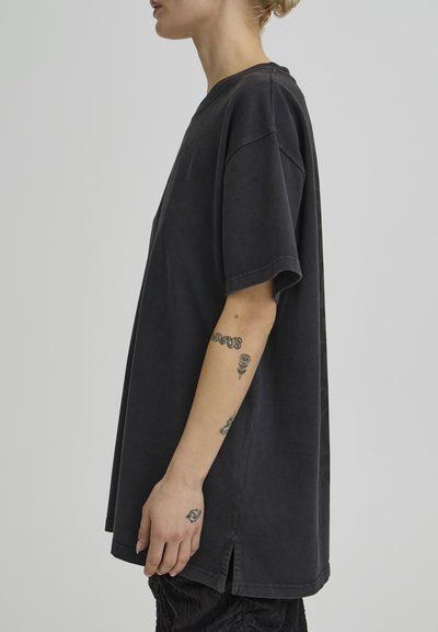 Személy túlméretezett fekete, rövid ujjú ingben, oldalán hasításokkal, előkarján és kezén tetoválások láthatók, egyszerű háttér előtt �áll.