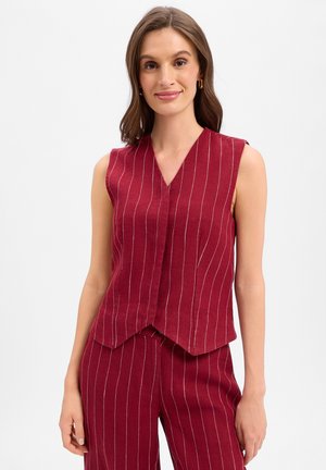 Femme portant un haut sans manches à fines rayures rouge foncé et un pantalon assorti, debout avec un léger sourire devant un fond uni.
