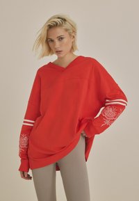 Červený oversized svetr s výstřihem do V, dlouhými rukávy s bílými pruhy a vyšitými květinovými vzory. Měkká, hladká textura látky.