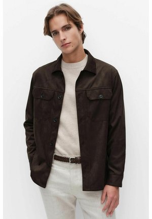 Jeune homme portant une veste boutonnée marron foncé sur un pull beige et un pantalon beige clair, debout devant un fond blanc uni.