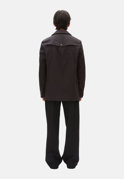 Schwarze taillierte Jacke mit breiten Revers, einem einzelnen Knopfverschluss auf der Rückseite und strukturierten Schultern. Kombiniert mit lockeren schwarzen Hosen.