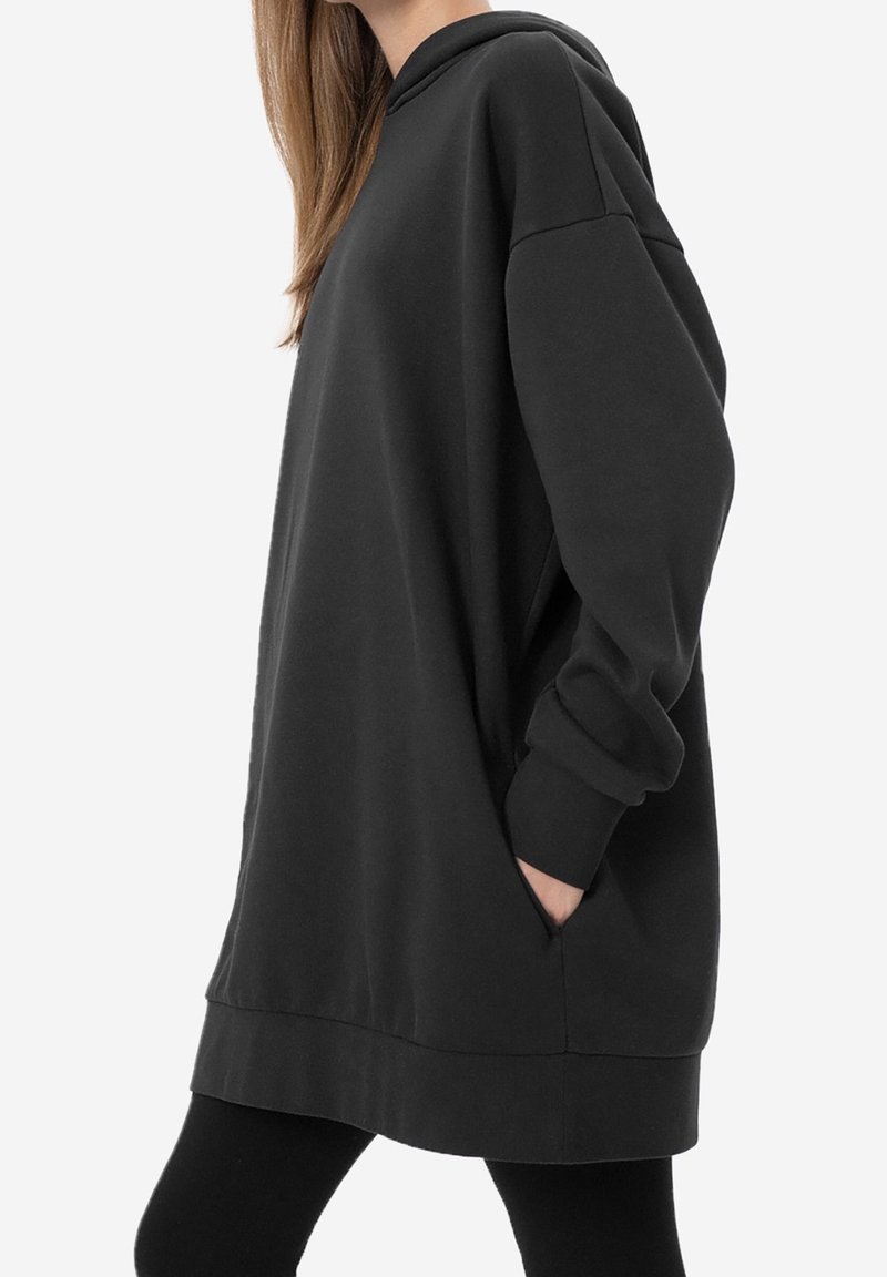 Femme portant un sweat à capuche noir long avec les mains dans les poches, associée à des leggings noirs, vue de profil.