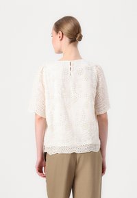 Spetsblus i krämfärgad med blommönster och scalloped fåll. Har kort ärm och knappstängning i nacken bak.