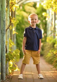 Granatowa koszulka polo z krótkim rękawem i guzikami, zestawiona z żółtymi szortami. Białe sneakersy dopełniają stylizację. W tle bujna zieleń.