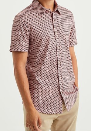 Mann trägt kurzärmeliges Hemd mit pinkem, schwarzem und weißem geometrischem Muster, das in beigefarbene Hose gesteckt ist.