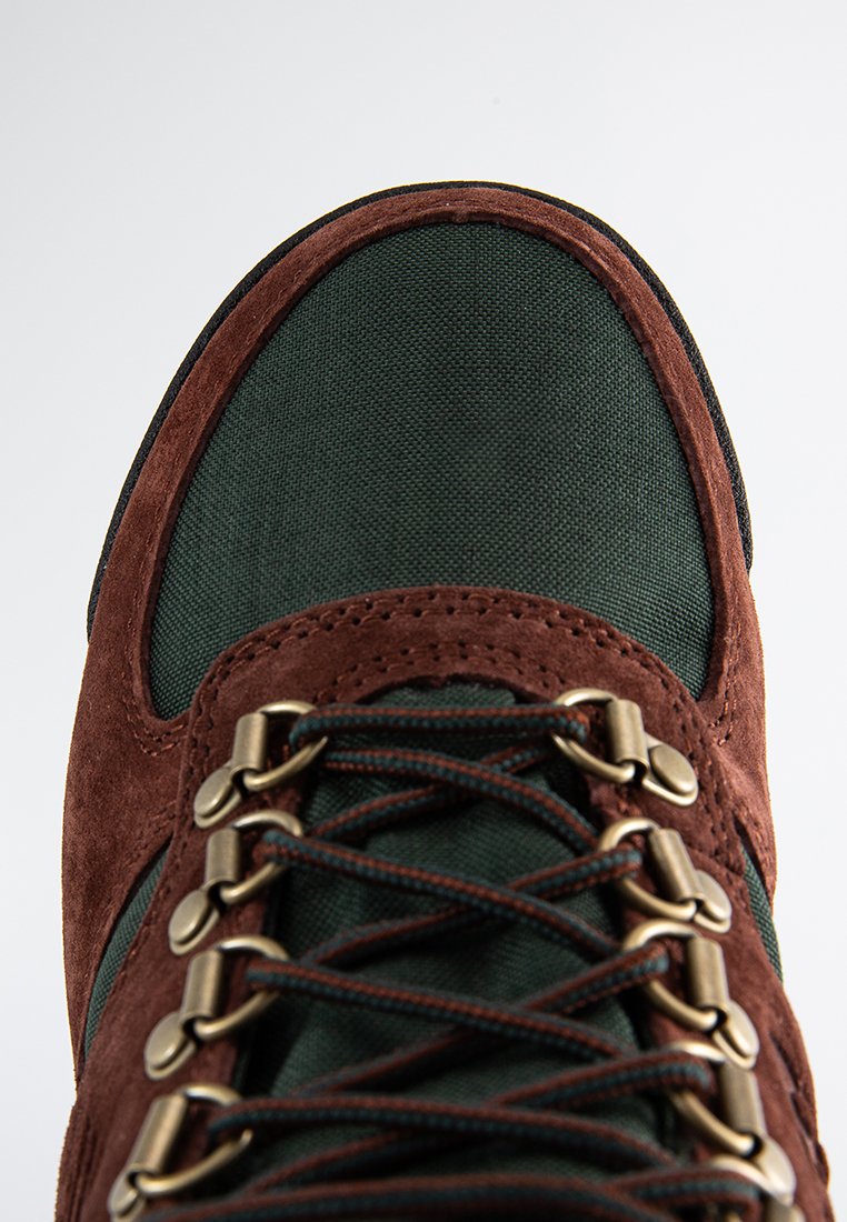 Chaussure en suède marron et tissu vert dotée d'œillets en métal, d'une languette texturée et de lacets marron. Le design est robuste et résistant.