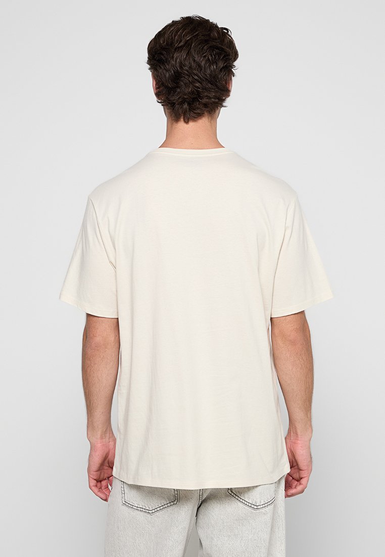 Camiseta de algodón beige con un ajuste relajado, mangas cortas y un escote redondeado. El dobladillo es ligeramente más largo en la parte posterior.