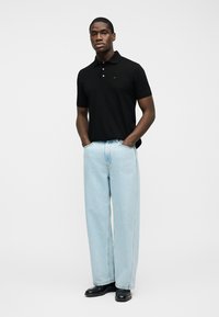 Άνδρας που φοράει μαύρο πόλο πουκάμισο, ανοιχτό μπλε τζιν παντελόνι με φαρδιά μπατζάκια και μαύρα παπούτσια, στέκεται με τα χέρια στις τσέπες μπροστά σε απλή φόντο.