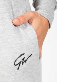 Gray sweatshirt fabric with a smooth texture, featuring a black signature logo on the pocket and a hand resting inside the pocket.