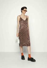 Mulher vestindo um vestido slip de estampado animal castanho, meias laranja, sapatos loafers pretos, óculos de sol castanhos, segurando uma bolsa branca, de pé contra um fundo branco liso.