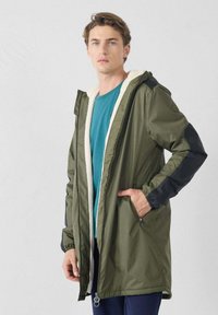 Parka verde con forro de piel sintética, detalles negros en las mangas, bolsillos laterales y cremallera en la parte inferior. Corte informal, diseño de longitud media.