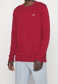 Czerwony sweter dziany z okrągłym dekoltem, ściągaczami na rękawach i u dołu, z małym logo jako akcentem. Połączony z jasnoniebieskimi dżinsami.