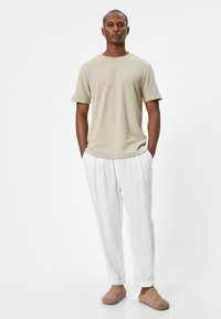 Maglietta a maniche corte color khaki abbinata a pantaloni bianchi con pieghe e scarpe slip-on beige, esposta su uno sfondo neutro.