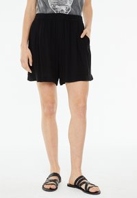 Pantaloni corti neri a pieghe con vita elasticizzata e tasche laterali, realizzati in materiale leggero, abbinati a sandali neri con cinturino.