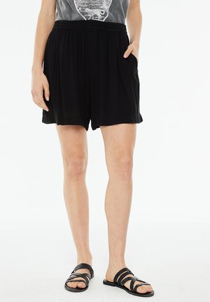Pantaloni corti neri a pieghe con vita elasticizzata e tasche laterali, realizzati in materiale leggero, abbinati a sandali neri con cinturino.