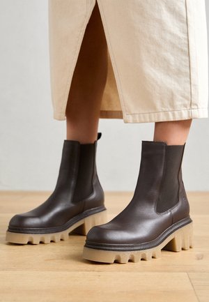 Bottes en cuir marron foncé montant jusqu'à la cheville, avec des semelles épaisses crantées beige, portées avec une jupe beige fendue à l'avant.
