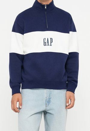 Mand iført en navyblå og hvid sweatshirt med kvart-lynlås med "GAP"-logo på brystet og lyseblå jeans, stående foran en ensfarvet baggrund.