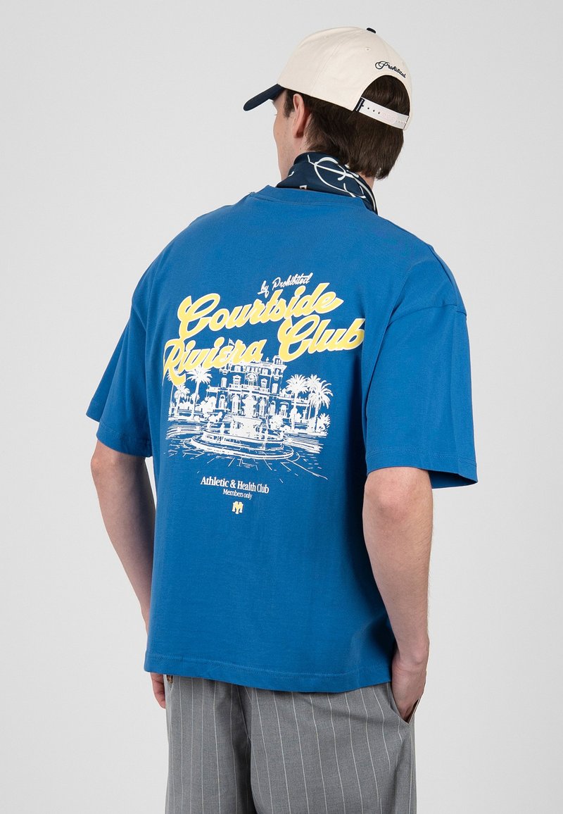 Homme portant un t-shirt bleu avec le texte jaune et blanc "Courtside Riviera Club" et un graphique de bâtiment, pantalon gris à fines rayures, casquette blanche et écharpe bleu marine.