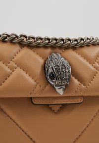 Prošívaná béžová kožená taška s tlustým stříbrným řetízkem a podrobným stříbrným uzávěrem ve tvaru ptáčí hlavy s černým drahokamovým okem.
