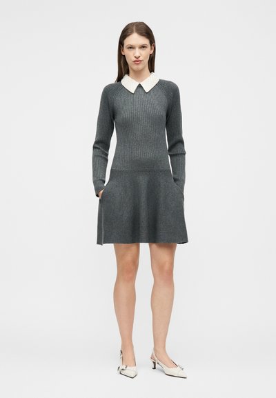 Femme debout portant une robe grise en maille côtelée à manches longues, col blanc texturé, poches latérales et chaussures blanches à talons bas avec bride arrière.