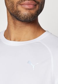 Camiseta atlética blanca hecha de una tela texturizada, con un logo de Puma de color azul claro, escote redondo y un detalle de costura sutil.