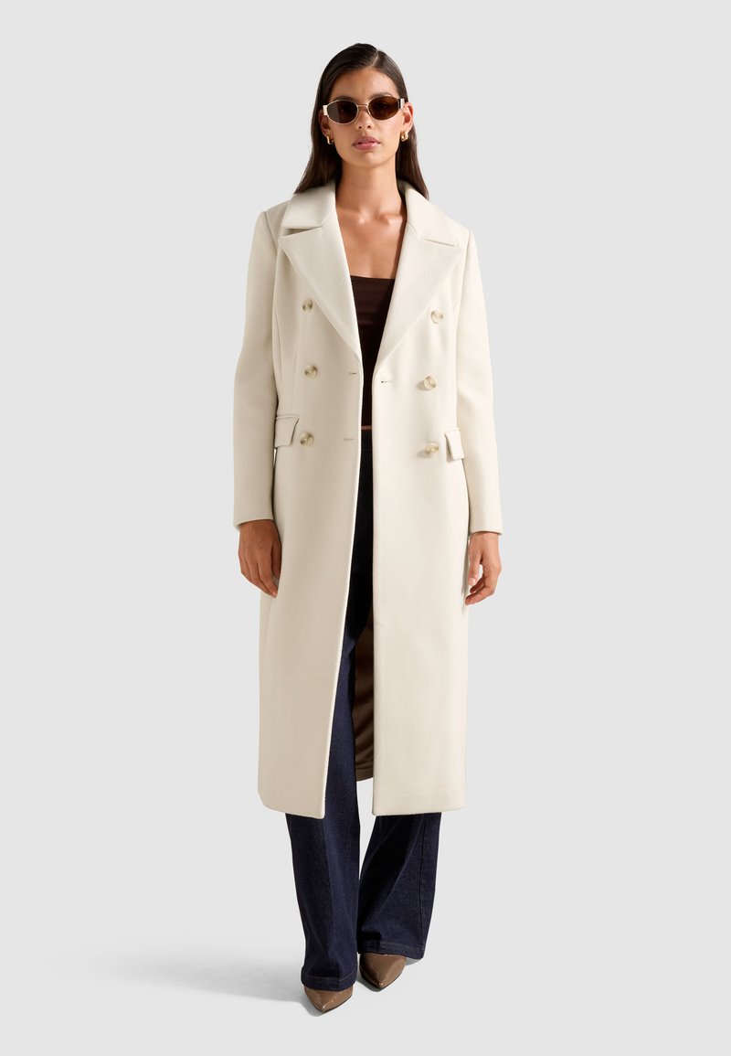 Cappotto lungo doppiopetto in crema, realizzato in materiale morbido, con revers strutturati e tasche laterali. Indossato sopra un top marrone e jeans scuri.